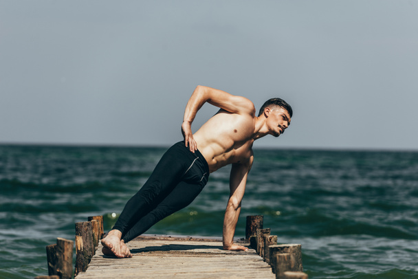 Sportlicher, hemdsloser Mann macht Seitenplanke auf Holzsteg - Foto, Bild