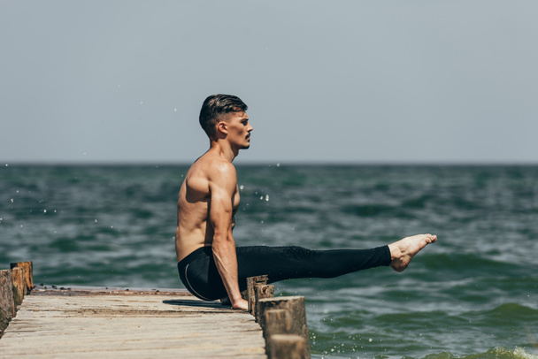 schöner hemdloser Mann praktiziert Yoga und macht Armbalance auf Pier - Foto, Bild