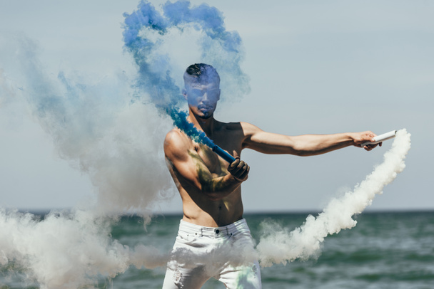 attractive man holding blue and white smoke sticks in front of sea - Foto, Imagem