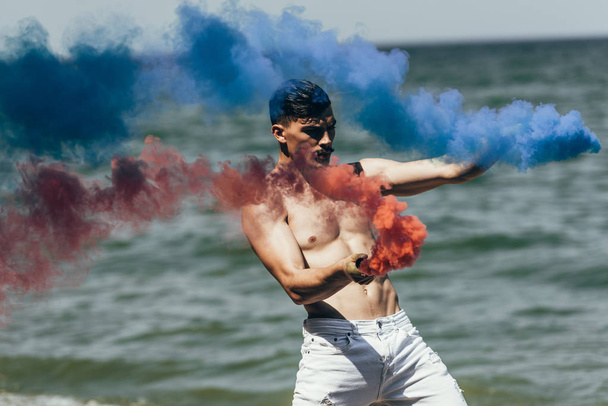 shirtless man dancing with red and blue smoke sticks in front of ocean view - Photo, Image