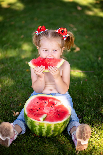 Μικρό κορίτσι τρώει καρπούζι στο γρασίδι, το καλοκαίρι. κοπέλα ντυμένη στα μπλε τζιν συνεδρίαση για την πράσινη χλόη τρώγοντας νόστιμα καρπούζι - Φωτογραφία, εικόνα