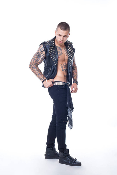 A young man with tattoos in a black leather vest with rhinestones and black jeans with a shiny belt is shod in shiny colors on a white background - Photo, Image