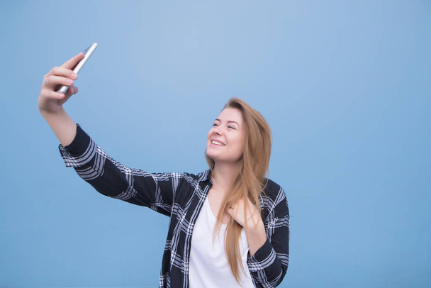 The happy girl in casual clothes is on a blue background and take selfie on a smartphone. Smiling girl poses on frontal camera smartphone - Zdjęcie, obraz