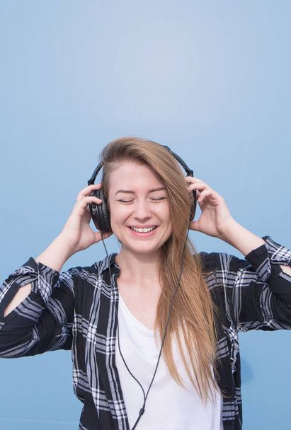 Happy girl in the casual clothing listens to music in cigarette holders with her eyes closed on a blue background. Student is listening to music isolated on a blue background. Copyspace - Fotó, kép