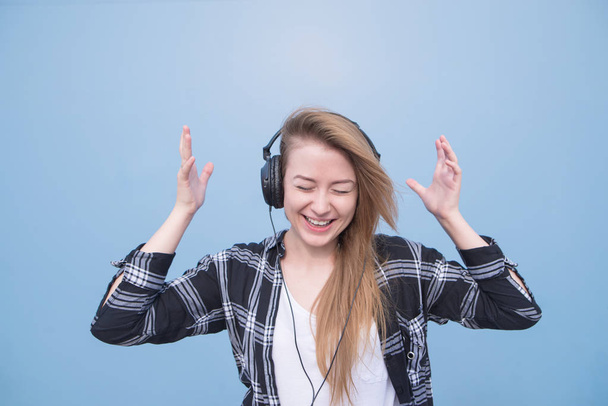 Positive girl in casual clothing listens to music in headphones, isolated on a blue background. Portrait of a happy woman listening to music on a blue background - Photo, Image