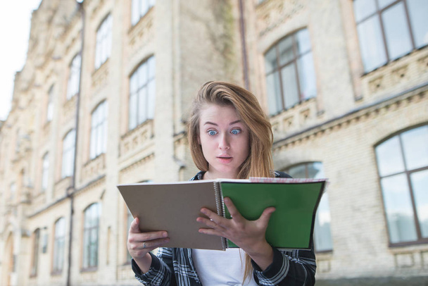 Hauska emotionaalinen tyttö opiskelija katsoo kirjaa hämmästyneenä yliopiston rakennuksen taustasta. Muotokuva opiskelijasta, joka näyttää yllättyneeltä kädessään olevasta kirjasta
. - Valokuva, kuva