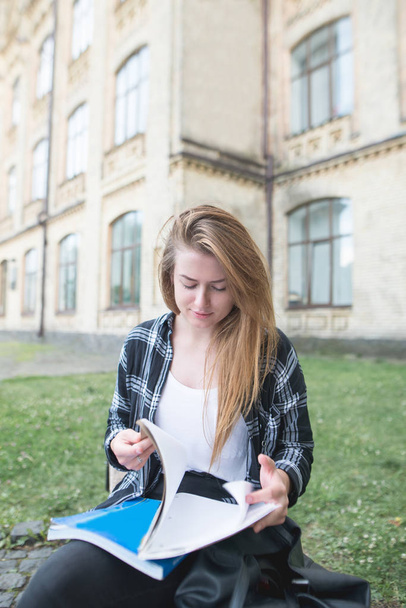 Young beautiful woman sitting on a bench on the background of the university and turning pages of the book. A girl reads on campus near a college building. Student Concept. - Photo, image