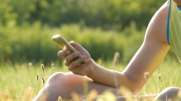 Mladý muž se dívá na smartphone a něco vytiskne na zelené trávě na pozadí krásné letní přírody - Záběry, video