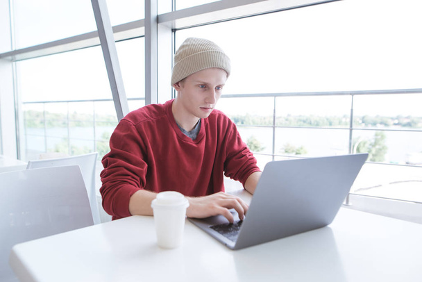 Aantrekkelijke jonge zakenman in casual kleding werkt op een laptop in de buurt van het venster in een stijlvol licht café. Jonge stijlvolle freelancer werkt op een laptop aan de tafel in de buurt van het panoramisch venster. - Foto, afbeelding