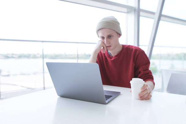 El joven freelancer se sienta en un café con un vaso de café en la mano y trabaja en un portátil. Retrato de un joven hombre de negocios con ropa casual trabajando en un portátil durante el almuerzo. Concéntrate en el portátil
 - Foto, imagen