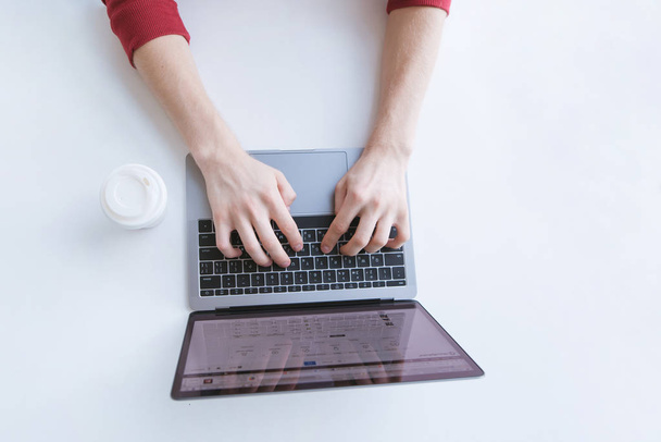 Man de tekst te typen op het toetsenbord van de laptop en een wit glas koffie op een lichte achtergrond. Werken op de werkplek. Bovenaanzicht. Copyspace - Foto, afbeelding