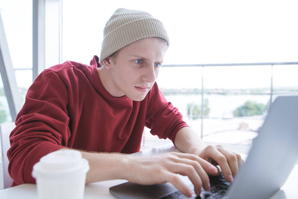 Poważne, młody człowiek w zwykłe ubrania, siedział przy stole i wpisywania tekstu na klawiaturze laptopa. Freelancer młodych działa na laptopie w kawiarni światła. Blogger pisze artykuł na komputerze - Zdjęcie, obraz