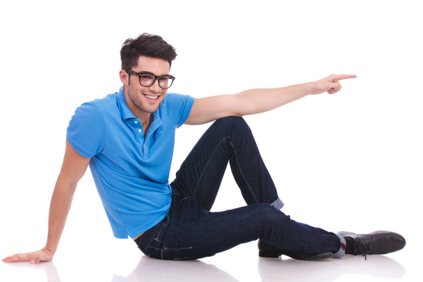 happy seated casual man is pointing to side while lying down on the floor on white background - Photo, Image