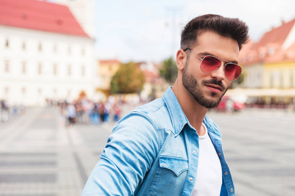 portrait of casual man with sunglasses standing outside in the city. He is wearing a blue denim shirt - Photo, Image