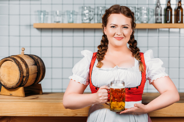 smiling oktoberfest bartender in traditional bavarian dress showing mug of light beer near bar counter - Foto, Imagem
