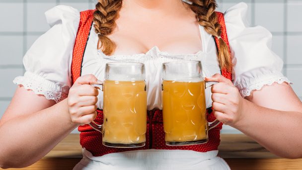 cropped image of oktoberfest waitress in traditional bavarian dress holding mugs of light beer with foam near bar counter - Photo, Image