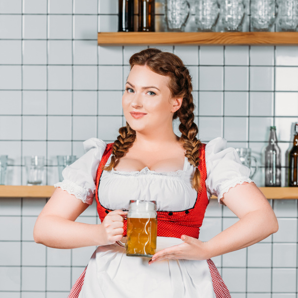 beautiful oktoberfest waitress in traditional bavarian dress showing at mug of light beer near bar counter - Photo, Image