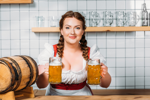 smiling oktoberfest waitress in traditional bavarian dress giving two mugs of light beer at bar counter  - Foto, imagen