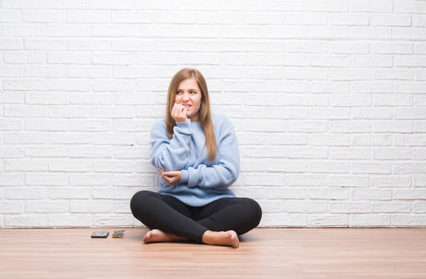 Nuori aikuinen nainen istuu lattialla syksyllä valkoisen tiiliseinän yli näyttäen stressaantuneelta ja hermostuneelta kädet suulla pureen kynsiä. Ahdistuneisuusongelma
. - Valokuva, kuva