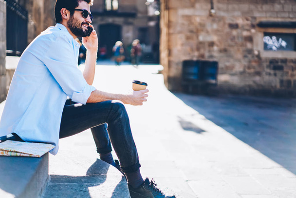 İşleci ile mobil yararları hakkında elinde lezzetli kahve ile sokakta oturan Smartphone'da konuşan sakallı genç adam. Neşeli turist üzerinde hücresel iletişim kurulurken gülüyor kırpılmış görüntü - Fotoğraf, Görsel
