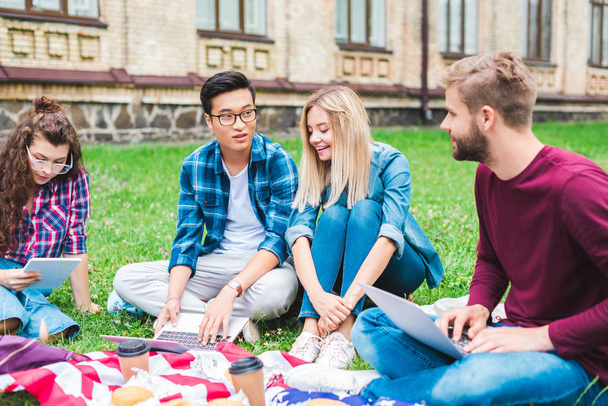 multicultural students with digital devices, coffee to go and american flag resting on green grass  - 写真・画像
