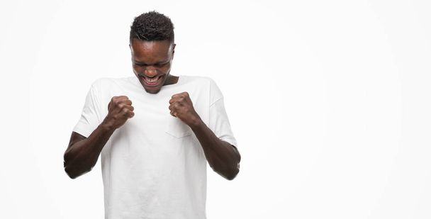 Młody człowiek african american ubrani na biało, t-shirt podekscytowany na sukces z ramionami wywoływane, docenianie zwycięstwa uśmiechnięte. Koncepcja zwycięzca. - Zdjęcie, obraz