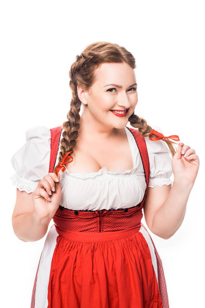 smiling oktoberfest waitress in traditional bavarian dress holding pigtails isolated on white background - Photo, Image