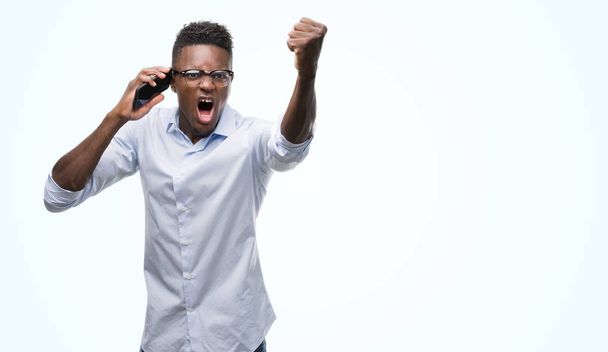 Fiatal afroamerikai férfi használ smartphone bosszús és csalódott kiabálás, a harag, őrült, és ordítás emelt kézzel, harag koncepció - Fotó, kép