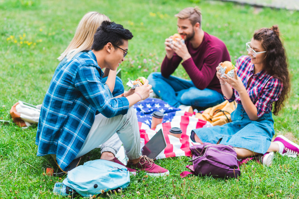estudiantes multirraciales con hamburguesas y bandera americana descansando en el parque
  - Foto, imagen