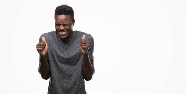 Jeune homme afro-américain portant un t-shirt gris excité pour le succès avec les bras levés célébrant la victoire en souriant. Concept gagnant
. - Photo, image