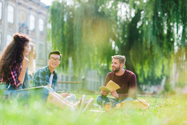 Lächelnde multirassische Studenten ruhen sich im Park mit Universität im Hintergrund aus - Foto, Bild