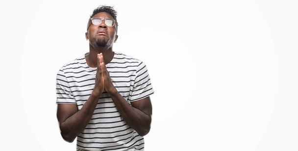 Młody człowiek african american, okularów i granatowy t-shirt żebractwa i modląc się z rąk wraz z nadzieją wyraz twarzy bardzo emocjonalne i martwi. Prosząc o przebaczenie. Koncepcja religii. - Zdjęcie, obraz