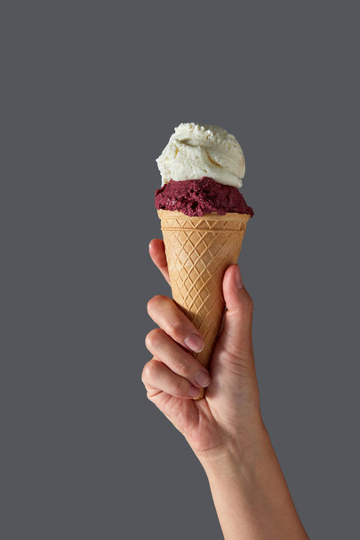 A female hand holds red berry gelato and white milk ice cream in a waffle cone on a gray background. Summer concept with copy space. - Φωτογραφία, εικόνα