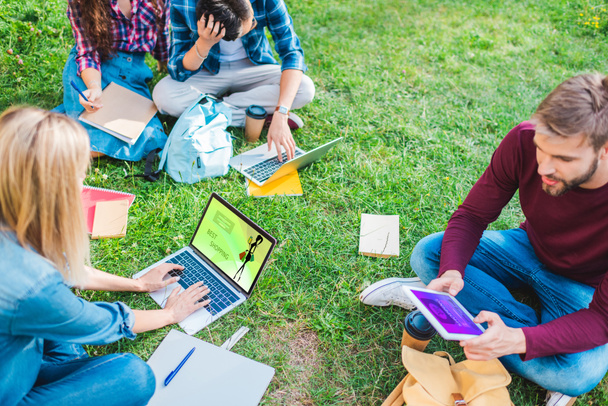 partial view of multiethnic students with notebooks and digital devices sitting on green grass in park - Photo, Image