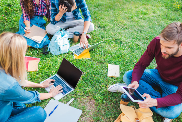 partial view of multiethnic students with notebooks and digital devices sitting on green grass in park - Foto, Bild