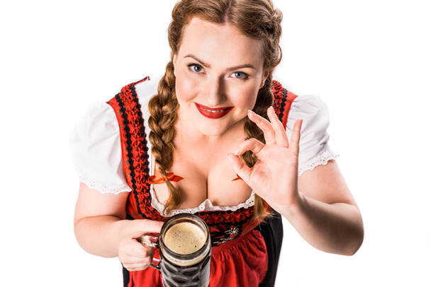 Hochwinkelaufnahme der Oktoberfestkellnerin in traditioneller bayerischer Tracht, die einen Becher dunkles Bier in der Hand hält und auf weißem Hintergrund vereinzelt OK-Zeichen zeigt - Foto, Bild