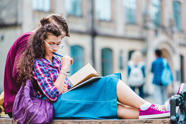 partial view of young woman with book and notebook leaning on boyfriend while studying on wooden bench in park - Photo, Image