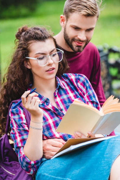 portrait of young woman with book and notebook leaning on boyfriend while studying on wooden bench in park - Photo, Image