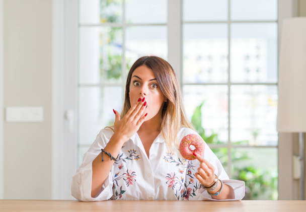 Young woman at home eating a doughnut cover mouth with hand shocked with shame for mistake, expression of fear, scared in silence, secret concept - Foto, Imagem