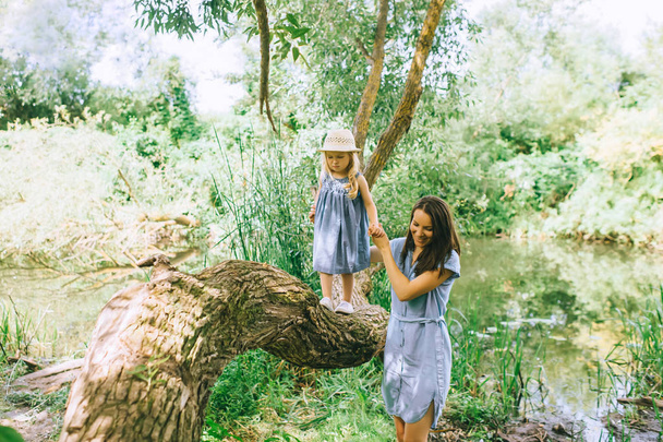 mother holding hands with daughter and helping her to walk on tree trunk, with lake on background - Photo, Image