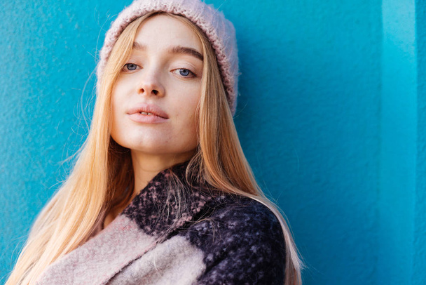 beautiful blue-eyed girl blonde in a warm hat posing against a blue wall background - Photo, Image