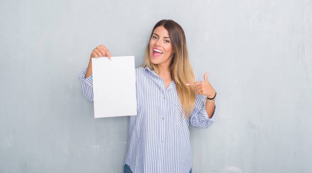 Молодая взрослая женщина на серой гранж-стене держит чистый лист бумаги с удивленным лицом, указывая пальцем на себя
 - Фото, изображение