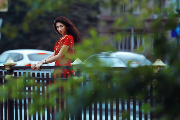 Uma linda garota bronzeada fica perto da cerca na ponte. Menina agradável em um fundo turvo da natureza. Menina bonito em um vestido vermelho. A vida cotidiana de uma bela jovem
. - Foto, Imagem