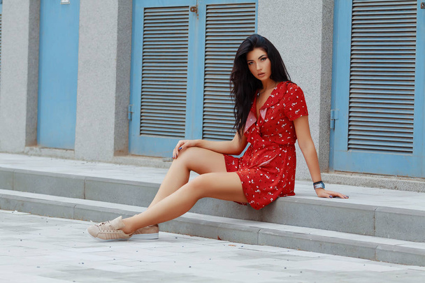 Νέοι μελαχρινή κοπέλα σαν να περιμένουν για κάποιον, που κάθονται στα σκαλοπάτια σε κοντό κόκκινο φόρεμα γυμνά πόδια, πλάγια όψη, γκρίζο τοίχο και μπλε πόρτα στο φόντο - Φωτογραφία, εικόνα