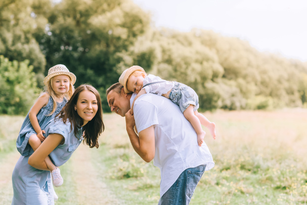 happy parents piggybacking son and daughter in straw hats on summer feild - Photo, Image