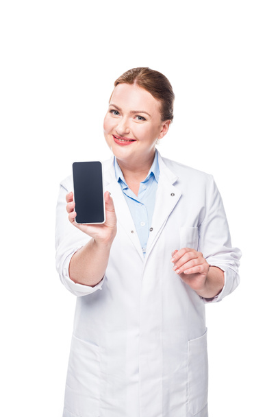 usmívající se lékařka ukazuje smartphone s prázdnou obrazovkou izolovaných na bílém pozadí - Fotografie, Obrázek