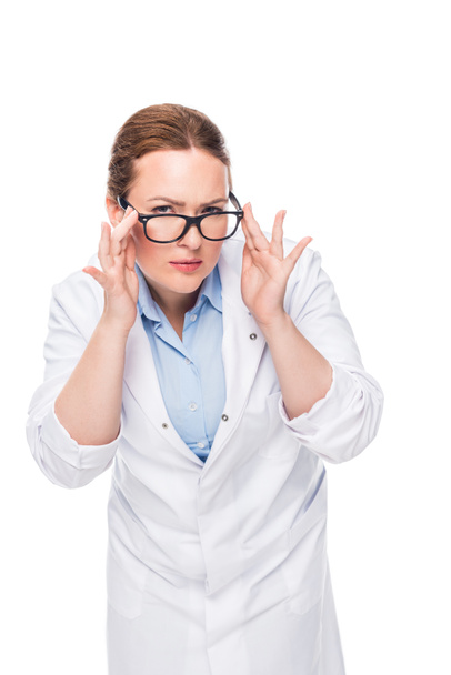 confident female doctor adjusting eyeglasses and looking at camera isolated on white background - Fotó, kép