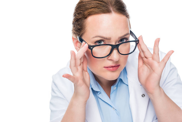 serious female doctor adjusting eyeglasses and looking at camera isolated on white background - Photo, Image