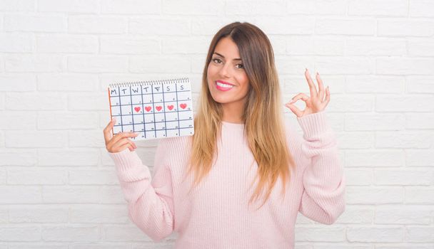 junge erwachsene Frau über weiße Backsteinmauer halten Zeitraum Kalender tun ok Zeichen mit den Fingern, ausgezeichnetes Symbol - Foto, Bild