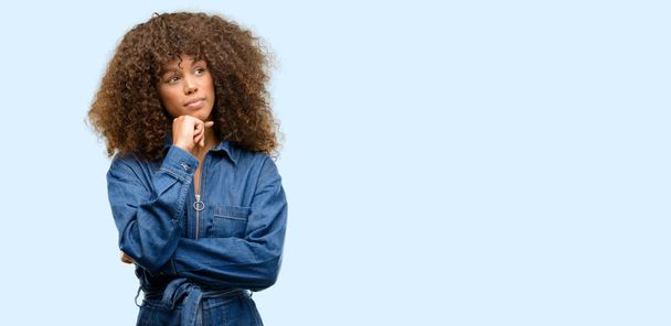 Африканская американка в синем комбинезоне думает и смотрит вверх, выражая сомнения и удивление
 - Фото, изображение